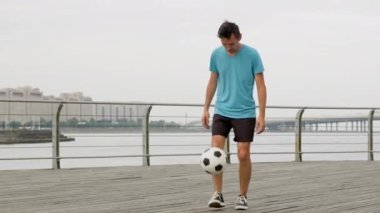 Man soccer player kicking bouncing a ball performing tricks on waterfront. Footballer practicing training outdoors juggling ball improving sport skills in slow motion. Sports football skills concept.