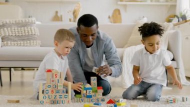 Black man is playing with children in living room of scrap building tower of wooden cubes. African american man has play with two children building tower of cubes sitting on floor of room.