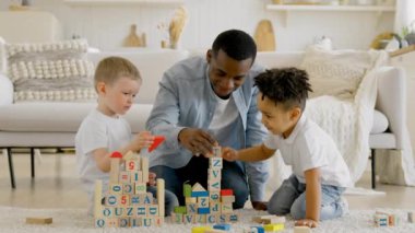 Black man is playing with children in living room of scrap building tower of wooden cubes. African american man has play with two children building tower of cubes sitting on floor of room.