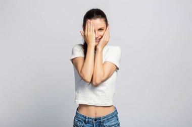 Neşeli genç ve çekici bir kadın yüzünün yarısını elleriyle saklıyor açık gri stüdyo arka planında.