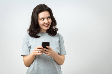 Neşeli, güzel, genç bir kız elinde bir cep telefonu tutuyor ve başka yere bakıyor, kopyalama alanı, gri arka planda izole edilmiş.