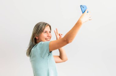 20 'li yaşlarda neşeli bir kadının portresi. Gri arka planda izole bir şekilde akıllı telefondan selfie çekerken gülüyor.