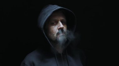 Siyah kapüşonlu bir adam burnundan sigara dumanı üflüyor. Karanlık atmosfer, siyah arkaplan.