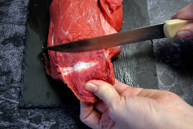 Knife in hand cutting fresh big beef meat piece on black background. Top view. Closeup