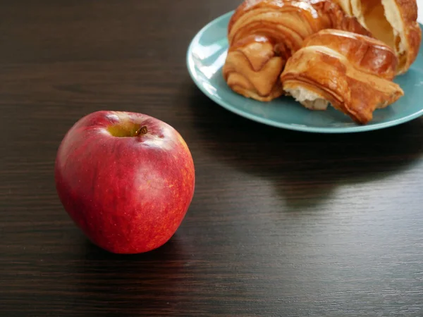 Red apple and croissants in plate on wooden background. Apple vs croissants, healthy food vs unhealthy food,  Closeup