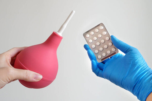 One hand holding enema and other hand holding pills in blister, enema vs pills for treatment concept, closeup