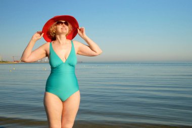Güneşli bir günde, açık havada, deniz sahilinde duran büyük kırmızı şapkalı ve retro mayo giyen orta yaşlı sarışın kadın.