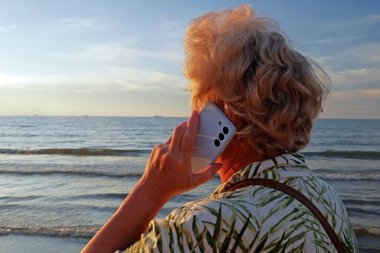 Sarışın, kıdemli bir kadın gün batımında, açık havada, denizde veya okyanus kıyısında akıllı telefonlardan konuşuyor.