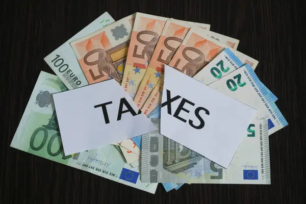 the cutting paper with inscription taxes lies on top of a large pile of euro money on a dark wooden table, top view, tax reduction concept