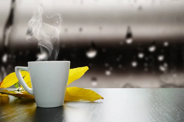 White cup of hot tea with smoke and autumn leaves on black wooden table with autumn rain drops on window glass, autumn mood concept, closeup