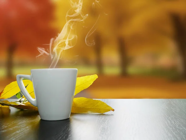 White cup of hot tea with smoke and autumn leaves on black wooden table with autumn landscape outside the window, autumn mood concept, closeup