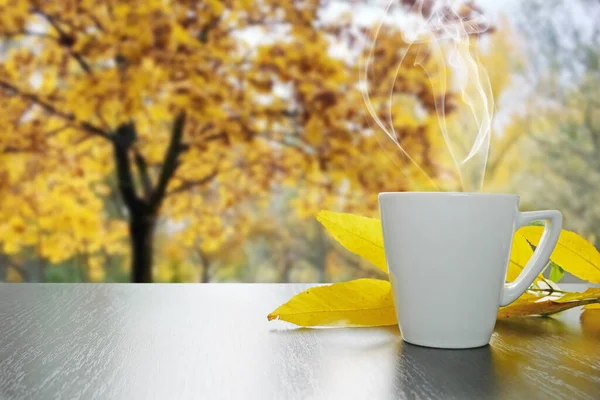 White cup of hot tea with smoke and autumn leaves on black wooden table with autumn landscape outside the window, autumn mood concept, closeup
