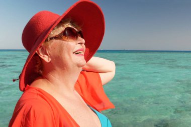 Mayo giymiş mutlu orta yaşlı kadın, büyük kırmızı güneş şapkası ve gözlük takan güneşli yaz gününde deniz arka planında kalan, tatil ve seyahat konsepti, açık havada