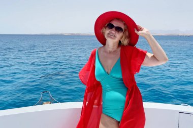 Orta yaşlı bir kadın tek parça mayo, kırmızı şapka, plaj pelerini ve güneş gözlüğü takıyor mavi deniz ve gökyüzü arka planında yatın keyfini çıkarıyor.