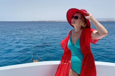 Orta yaşlı bir kadın tek parça mayo, kırmızı şapka, plaj pelerini ve güneş gözlüğü takıyor mavi deniz ve gökyüzü arka planında yatın keyfini çıkarıyor.