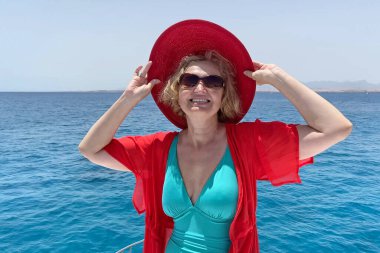 Orta yaşlı bir kadın tek parça mayo, kırmızı şapka, plaj pelerini ve güneş gözlüğü takıyor mavi deniz ve gökyüzü arka planında yatın keyfini çıkarıyor.