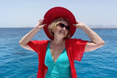 Orta yaşlı bir kadın tek parça mayo, kırmızı şapka, plaj pelerini ve güneş gözlüğü takıyor mavi deniz ve gökyüzü arka planında yatın keyfini çıkarıyor.