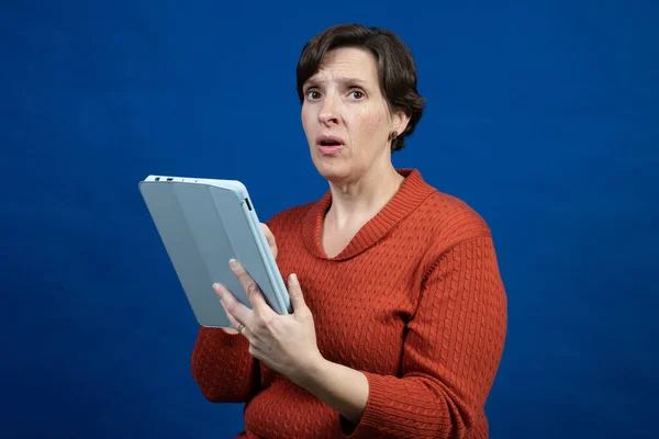 一个女人穿着橙色的寒冷天气毛衣 蓝色背景 看着相机 对她的电子阅读器做出了反应 — 图库照片