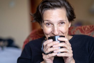 50 'li yaşlarda, siyah bluzlu bir kadın doğal ışık alan bir otelde bir fincan kahvenin tadını çıkarıyor..