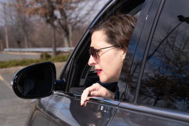 Sürücü koltuğunda oturan orta yaşlı bir kadın parlak bir kış gününde arabasının camından dışarı ruj sürerek bakıyor..