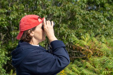 Middle-aged woman looking through binoculars wearing a bright red baseball cap on a sunny day. clipart