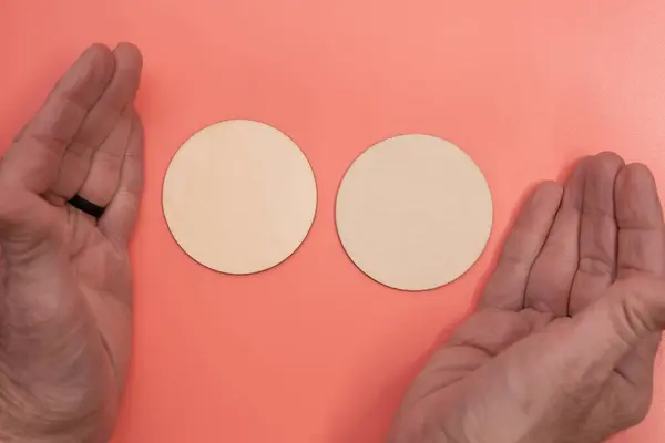 Two blank wooden disks with mans hands framing them on a light pink background with room for copy