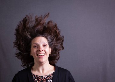 50 yaşında bir kadın saçlarını arkaya atar kamera hareketlerini gri bir arkaplanda dondurur.