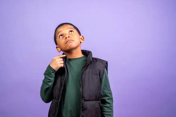 A young boy wearing a black vest and green shirt is looking up at the camera. The boy appears to be lost in thought, possibly contemplating something important. The image has a contemplative