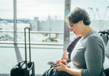 Bir kadın yanında bavulla bir sandalyede oturuyor. Telefonuna bakıyor ve bir deftere yazıyor.