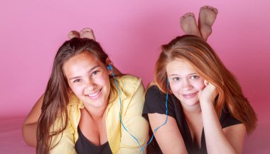 İki kız yerde yatıyor, biri sarı tişört giyiyor ve diğeri siyah gömlek giyiyor. İkisi de kulaklıkla müzik dinliyor.