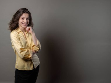 Sarı gömlekli bir kadın gri bir duvarın önünde duruyor. Kameraya ciddi bir ifadeyle bakıyor.