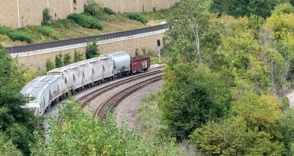 Tren arka planda yeşil bir tepe ile raylarda ilerliyor. Tren çok fazla yük taşıyor.