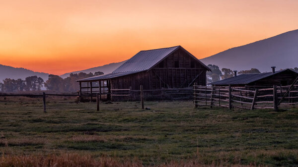 Большой, старый амбар сидит в поле с красивым закатом на заднем плане. Сцена спокойная и серенькая, амбар и окружающий пейзаж создают ощущение спокойствия и умиротворения