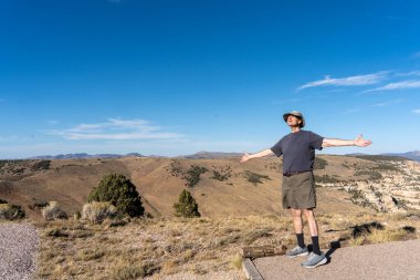 Şapkalı ve şortlu bir adam bir patikada dikiliyor ve bir dağa bakıyor. Gökyüzü mavi ve arka planda ağaçlar var.