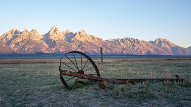 Old non-motorized farm equipment on a flat plain in front of the Teton Mountain range in Wyoming clipart