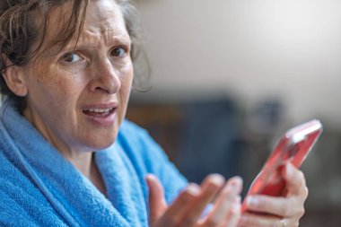 Bir kadın mavi bir bornoz giyerken telefonuna bakıyor. Dalgın bir ruh hali içinde.