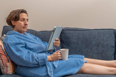 Mavi bornozlu bir kadın elinde bardak tutarken kanepede tablet okuyor. Kadın elinde sıcak bir içecekle koltukta rahatça oturduğu için rahatlama ve rahatlama kavramı.