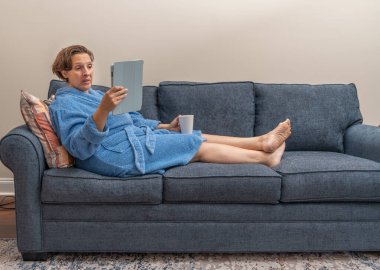 Mavi bornozlu bir kadın elinde bardak tutarken kanepede tablet okuyor. Kadın elinde sıcak bir içecekle koltukta rahatça oturduğu için rahatlama ve rahatlama kavramı.