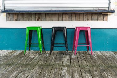 Tahta bir güvertede üç tabure oturuyor. Yeşil Siyah ve Pembe Tabureler