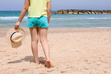 Şık şapkalı çocuk deniz kenarında kumda yürüyor.