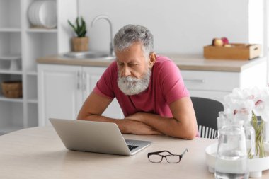 Mutfaktaki masada dizüstü bilgisayar kullanan yaşlı sakallı adam.