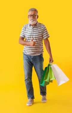 Alışveriş çantalı yaşlı adam sarı arka planda baş parmağını gösteriyor.