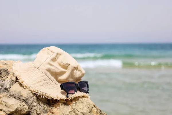 stock image Stylish bucket hat and sunglasses on rock near sea, closeup