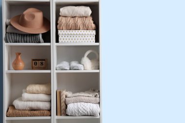 Mavi duvarın yanında kazaklı, şapkalı ve dekorlu raf ünitesi.