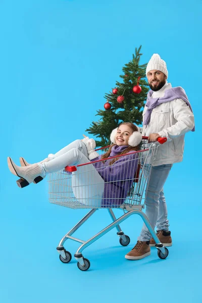 青を背景にクリスマスツリーとショッピングカートを持つ若いカップル — ストック写真