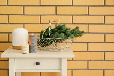 Sarı tuğla duvarın yanındaki masada Noel dalları ve mumları olan sepet.