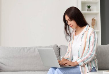 Genç Asyalı kadın evdeki kanepede dizüstü bilgisayar kullanıyor.