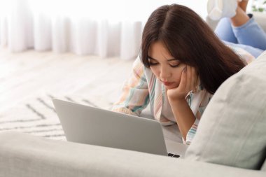 Genç Asyalı kadın evdeki kanepede dizüstü bilgisayar kullanıyor.