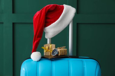 İçinde Noel Baba şapkası, Noel hediyeleri, pasaport ve pusula olan çanta yeşil duvarın yanında.