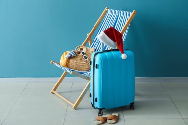 Noel Baba şapkalı bavul, güverte sandalyesi ve mavi duvarın yanında seyahat aksesuarları.